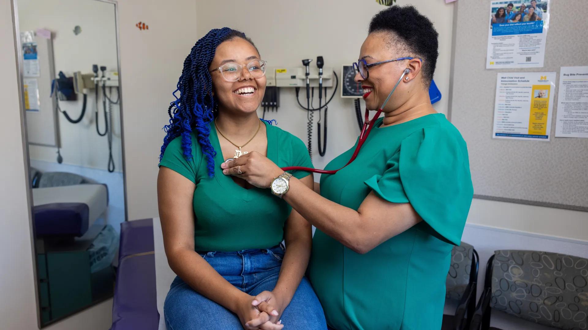 A youth with a teal shirt, blue hair, and glasses is examined by a woman provider with light skin, dark brown hair, brown eyes, and blue glasses.