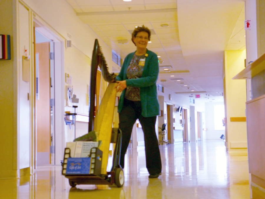 Woman wearing green sweater wheeling harp down hospital hallway