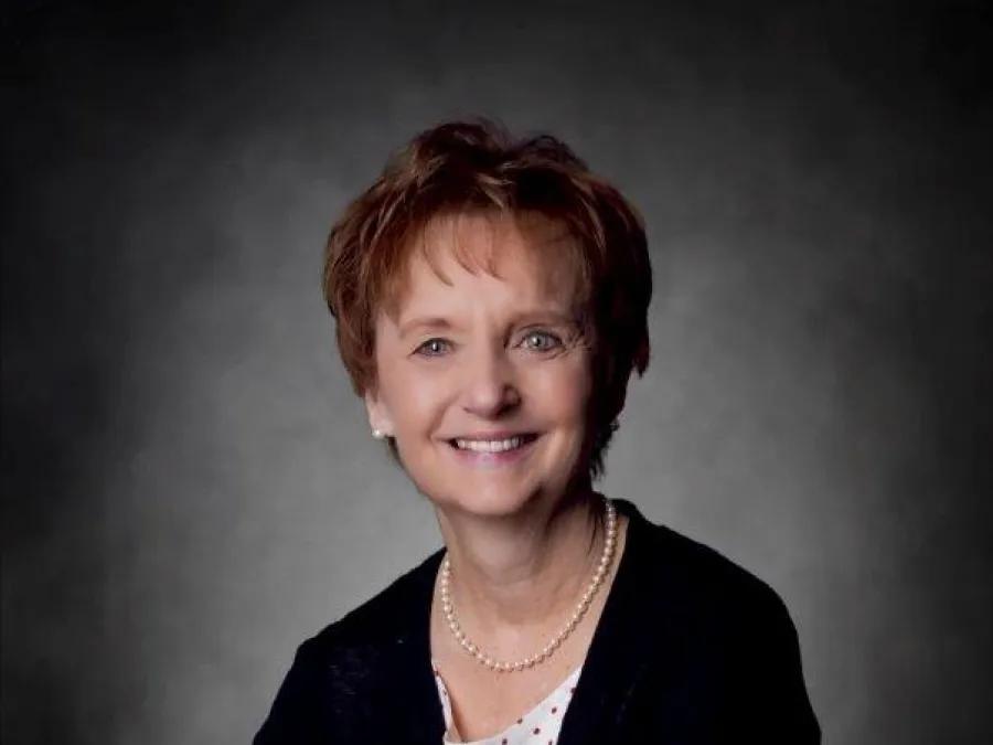 Smiling woman with reddish brown hair and hands folded wearing white blouse with black dots under black cardigan