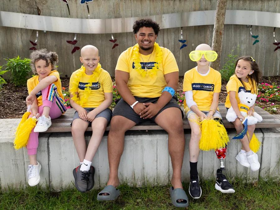Photo of an adult and four children seated on a concrete garden border. They are all smiling, wearing Block Out Cancer t-shirts, and some are holding fun items like pom poms and a plush bear.