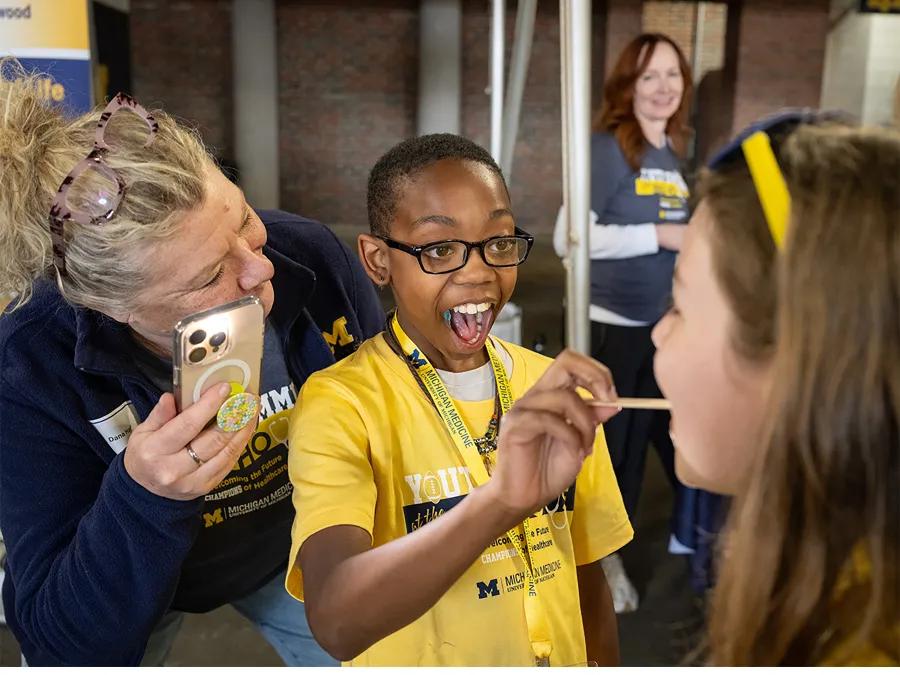 Young black boy with glasses and smiling with open mouth using tongue depressor next to smiling white woman with phone 