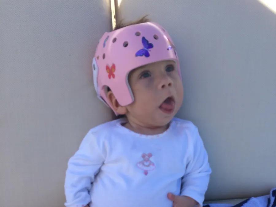 Baby sitting cross-legged on a patterned blanket, wearing a white onesie and a pink protective helmet decorated with butterflies and flowers.