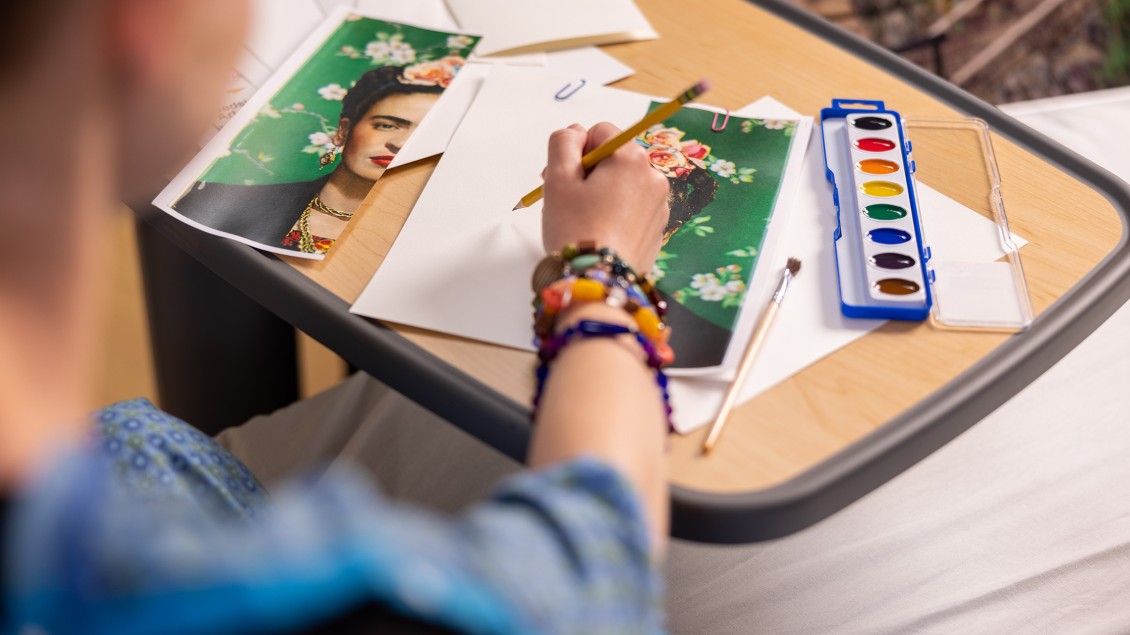 Woman's arm with colorful bracelets holding a pencil with art prints and paintbox on table