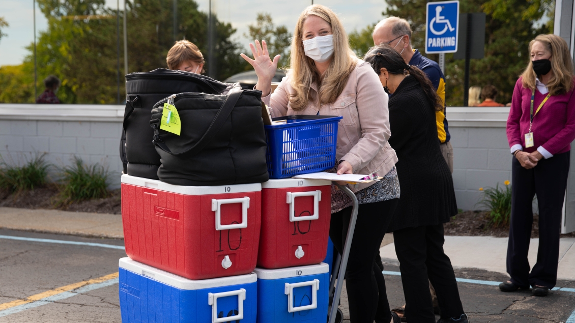 Woman with mask waving and pushing a cart stacked withred and blue coolers