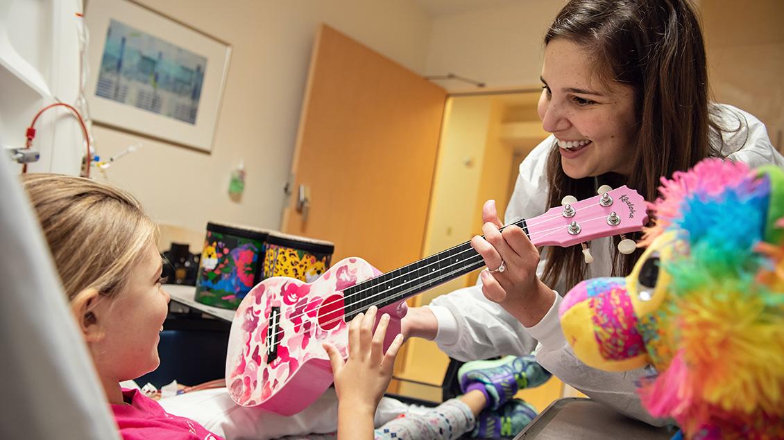 woman handing female pediatric patient laying in a hospital bed a pink guitar