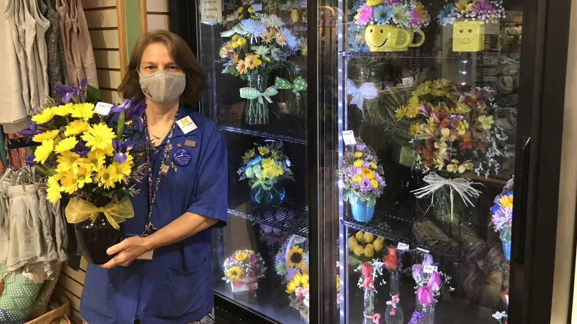 Volunteer standing with flower arrangement next to a glass-front refrigerator with flower arrangments inside
