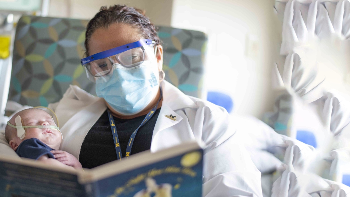 Nurse in goggles and a surgical mask reading to a sleeping baby patient