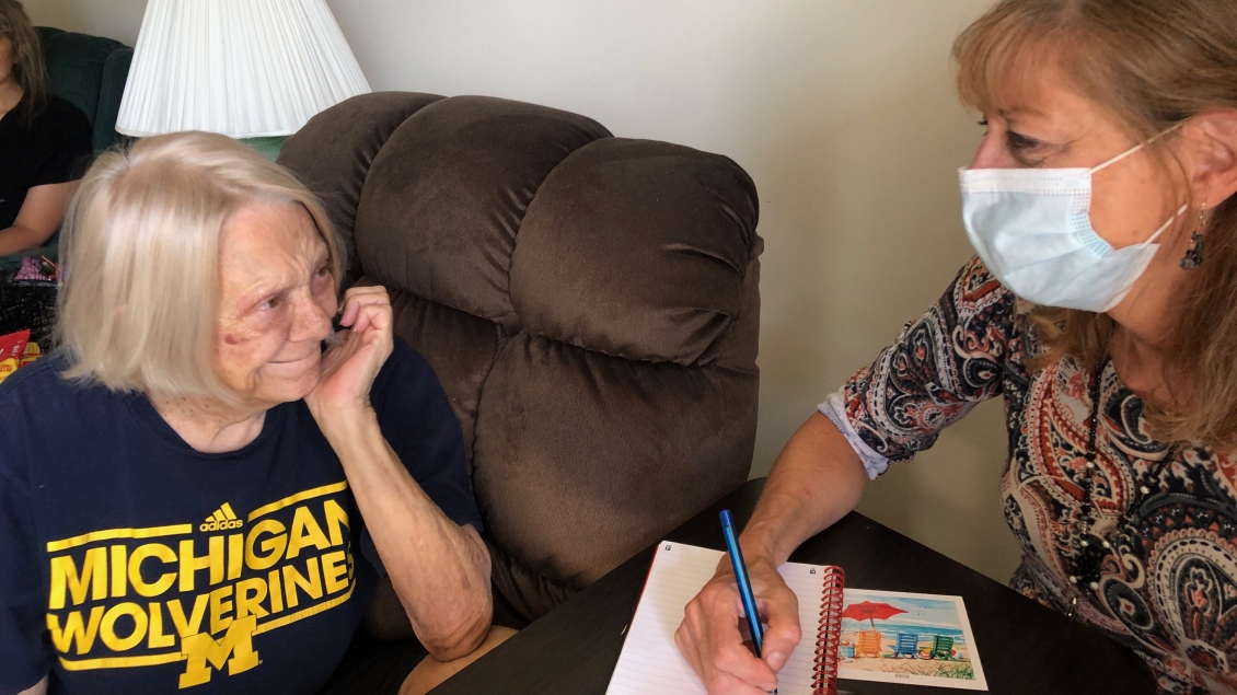 Elderly woman wearing Michigan Wolverine shirt sitting on sofa next to woman wearing mask and writing in notebook