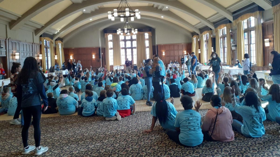Group of Neuroscience Graduate Program students sitting on the floor in a ballroom with matching light blue shirts listening to a presentation