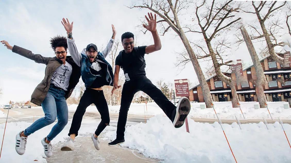 Three teens in mid-jump in an outside setting, hands raised and smiling, with snow and bare trees in the background
