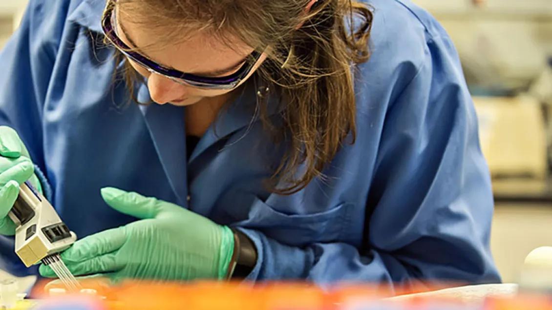 A woman wearing safety glasses, green gloves, and blue scrubs works with a piece of lab equipment.