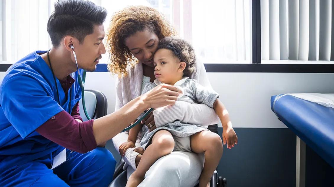A nurse examines a young child who is sitting on their mom's lap.