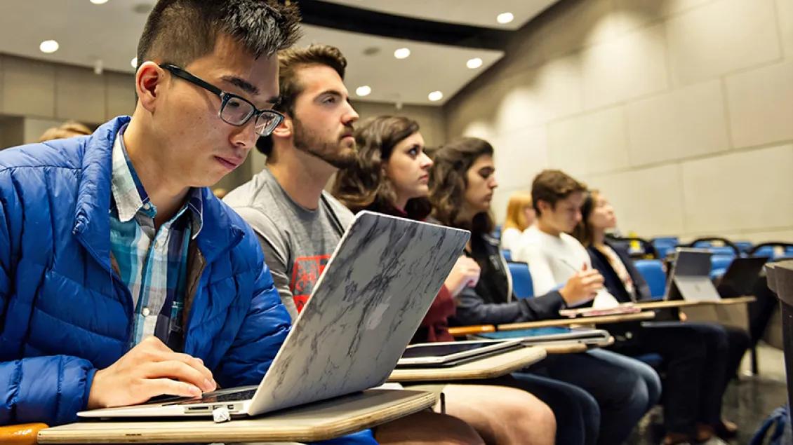 Medical students sit in a row in a classroom. The closest student is wearing glasses and a blue jacket and is working on a laptop.