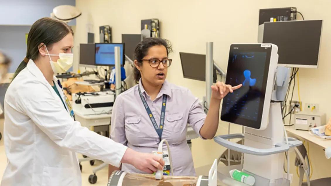 Two women work with a simulation program in a lab. One is holding the device and the other is pointing to a monitor.