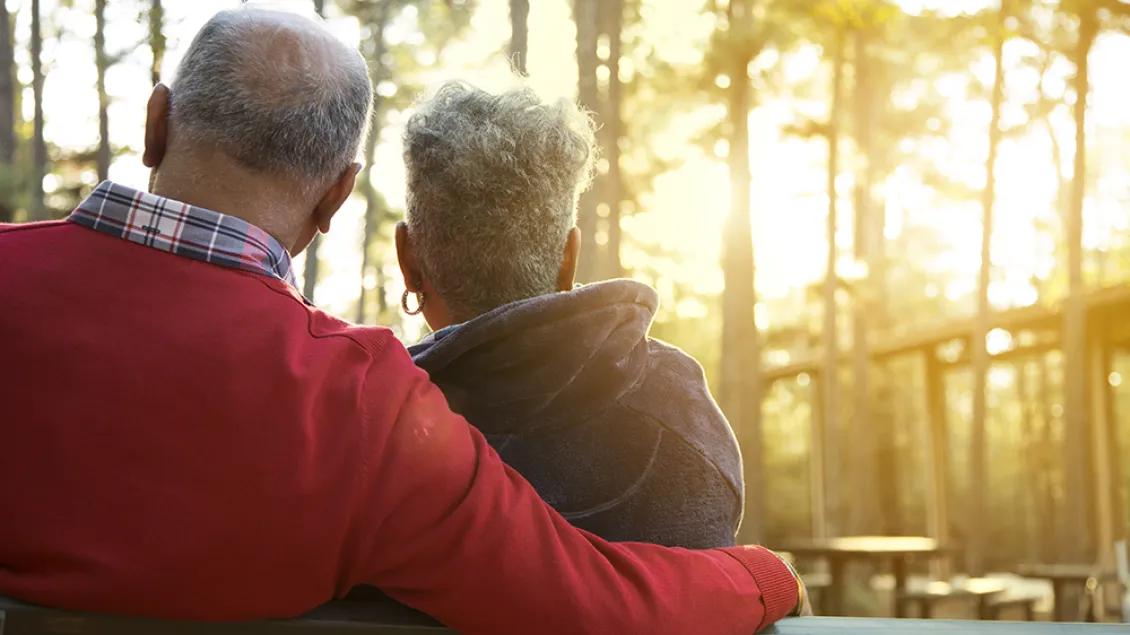 An older couple seated on a bench at the edge of the woods. They are facing away from the viewer.