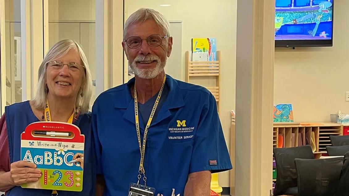 Two smiling volunteers stand with a mobile book cart filled with children's books.