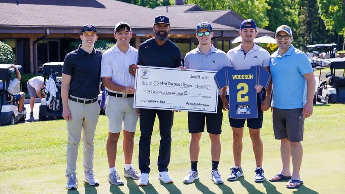 Charles Woodson holding an oversized check with five golfers. They are on a sunny golf course and other golfers and carts are visible behind them.