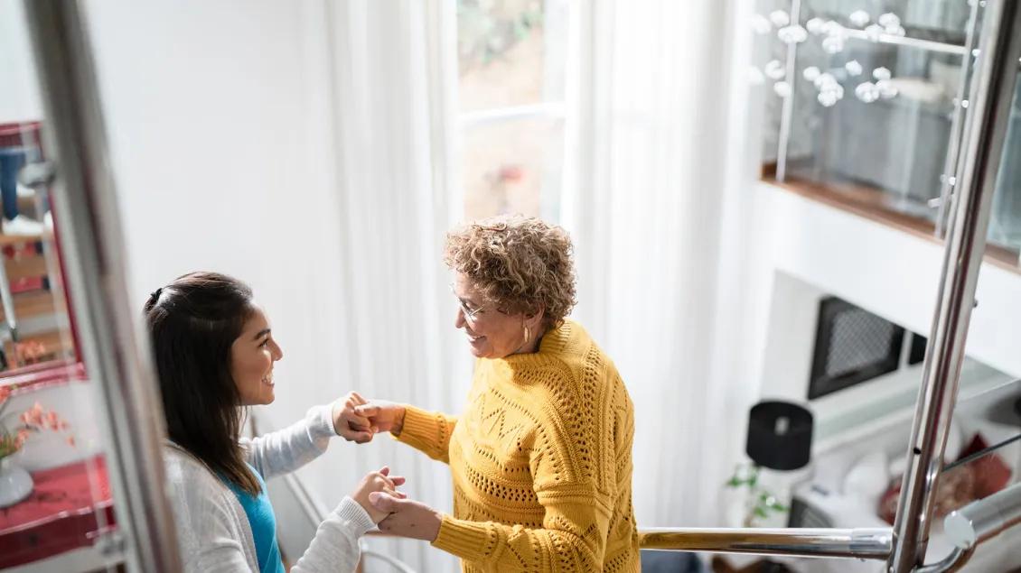 An older person and a younger person smiling and holding hands as they stand in a home.