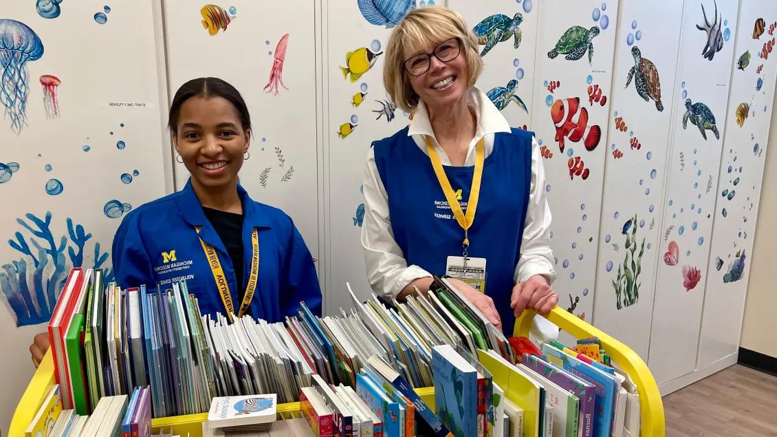 Two smiling Michigan Medicine Giving Library volunteers stand behind a rolling cart full of books for children of all ages.