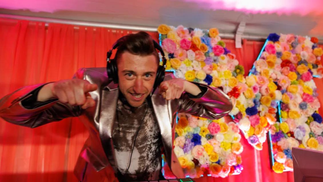 A person festively dressed in a metallic silver blazer and wearing a headset smiles and leans and points toward the viewer. There are dark pink curtains and a block M decorated with numerous roses in a variety of colors behind him.