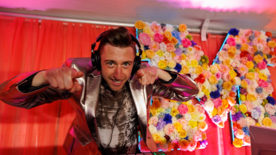 A person festively dressed in a metallic silver blazer and wearing a headset smiles and leans and points toward the viewer. There are dark pink curtains and a block M decorated with numerous roses in a variety of colors behind him.