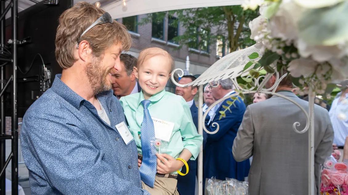An adult holds a young child at Event on Main. There is a crowd of people behind them and a bouquet of white flowers extends into the right side of the image.