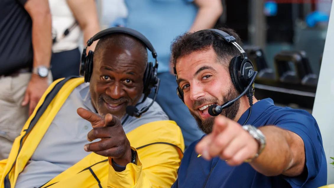 Two men wearing headsets smile and point towards the viewer. They are dressed in U-M colors with one wearing a yellow jacket and one wearing a blue shirt.