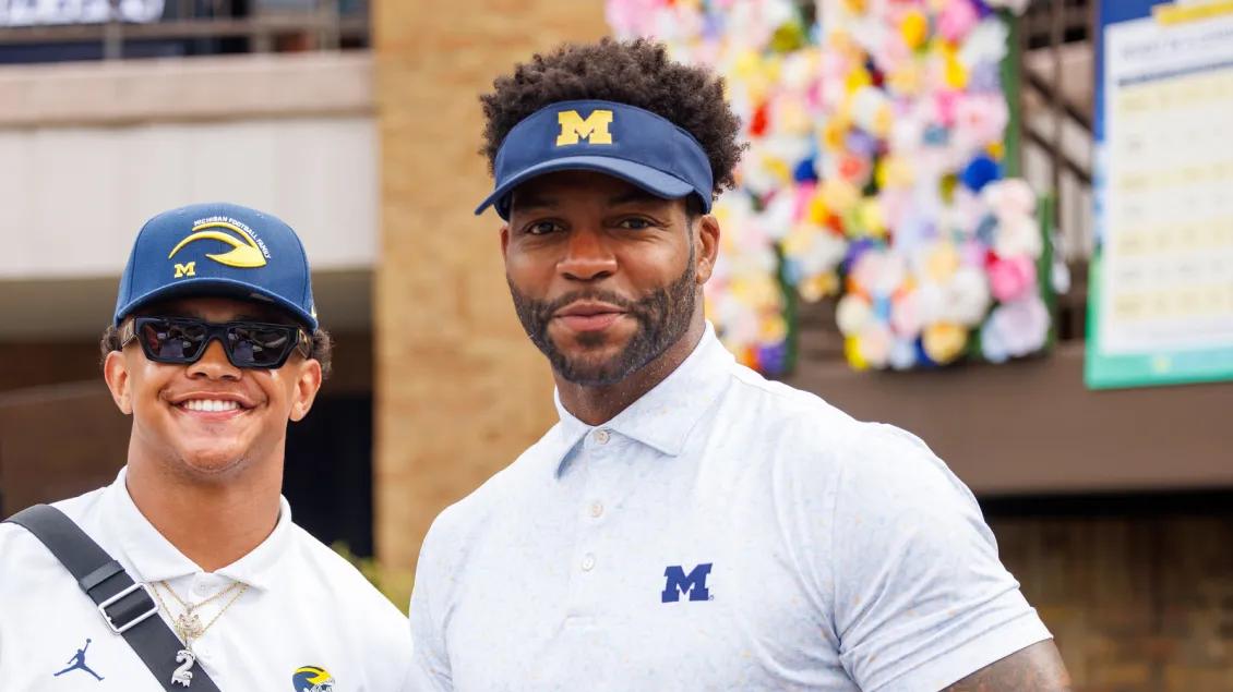 Portrait of two adults, one in a U-M baseball cap and one in a U-M visor, smiling toward the viewer. A block M covered in many colors of roses is behind them.