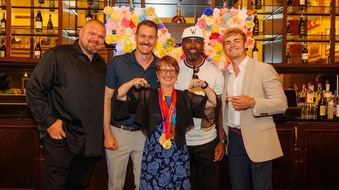Group photo including Dr. Donna Martin smiling and flexing her muscles as she wears two medals around her neck. Charles Woodson and three other people are standing behind her.
