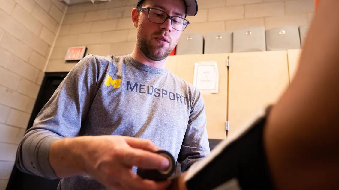 Close-up shot of a sports medicine professional wearing a U-M med sport t-shirt applying tape to an athlete's arm that is just visible in the foreground. 