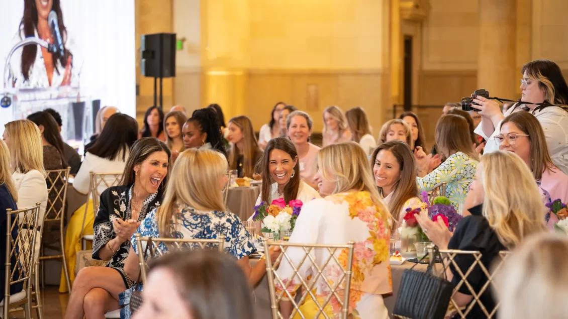 About 25 luncheon attendees seated around tables, smiling, and engaged in conversations. A photographer examines her camera on the right and there appears to be a presentation on a large screen to the left.