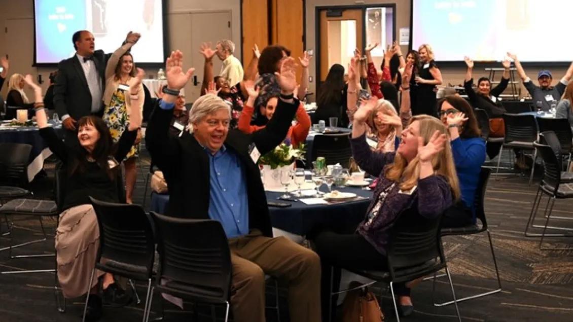 A group of about 20 adults seated around several round tables in a large conference room are smiling and raising their arms in what appears to be a celebratory manner.