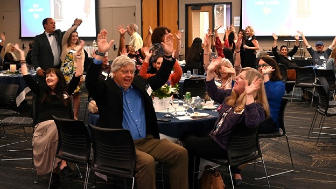 A group of about 20 adults seated around several round tables in a large conference room are smiling and raising their arms in what appears to be a celebratory manner.