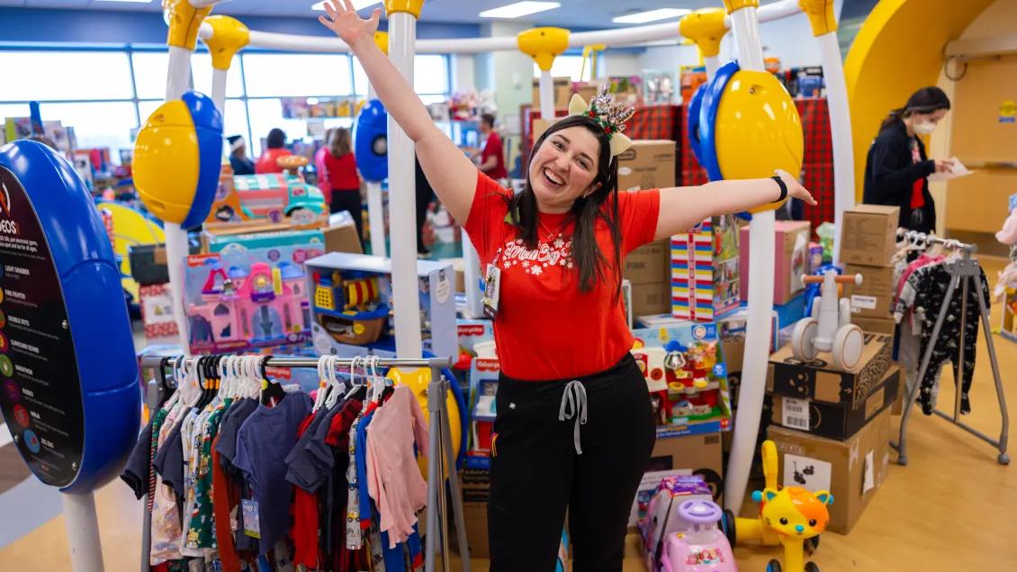 A staff or volunteer in a red t-shirt stands with her arms extended excitedly in front of a room full of kids' clothing and toys.