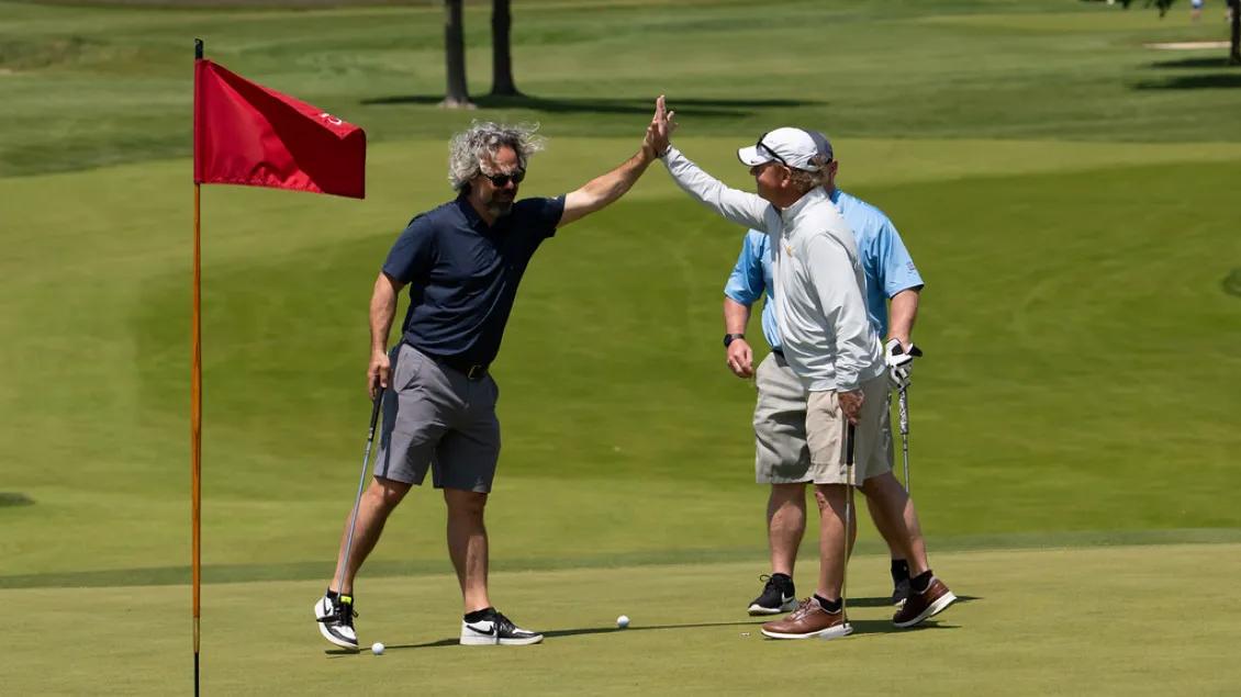 Two people standing on a golf green give each other a high five near a red flagstick, while a third person stands nearby holding a golf club. All are dressed in golf attire, and the scene is set on a sunny day with lush green grass in the background.