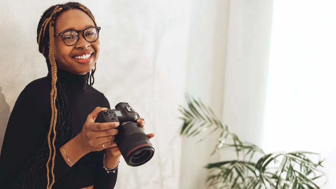 Young black woman with long braids and wearing glasses and a black turtleneck holding a camera and smiling