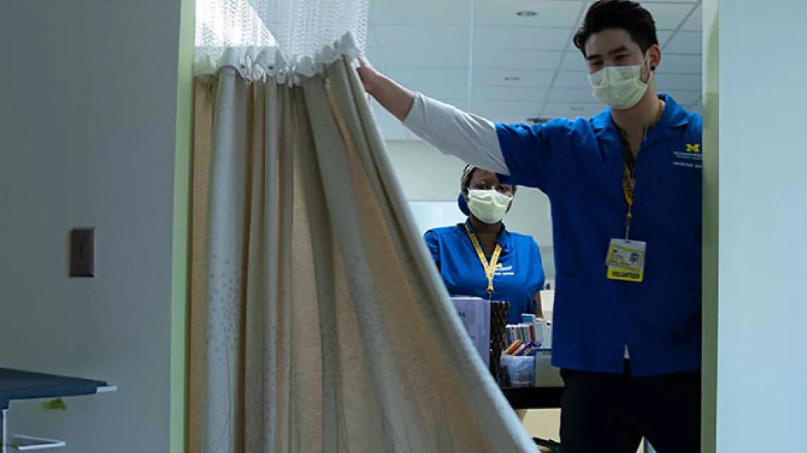 Two volunteers wearing blue shirts, yellow lanyards, and masks open a privacy curtain to enter a hospital bay.