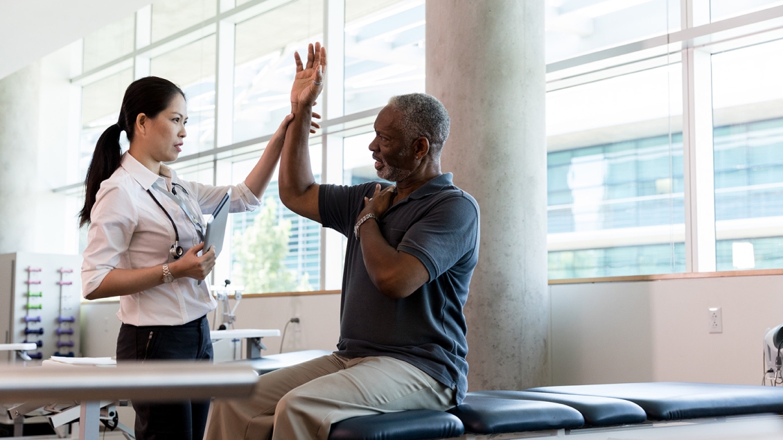 A medical professional stands next to a patient who is seated on an exam table. The patient is holding up his right arm, and the medical professional is loosely holding his wrist. They appear to be discussing his arm or shoulder.