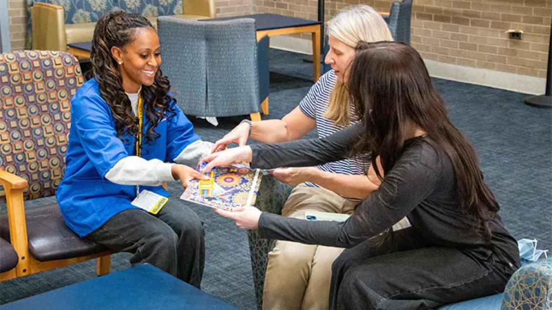 A smiling volunteer in a blue uniform hands a coloring book and crayons to two individuals sitting in a lounge area.