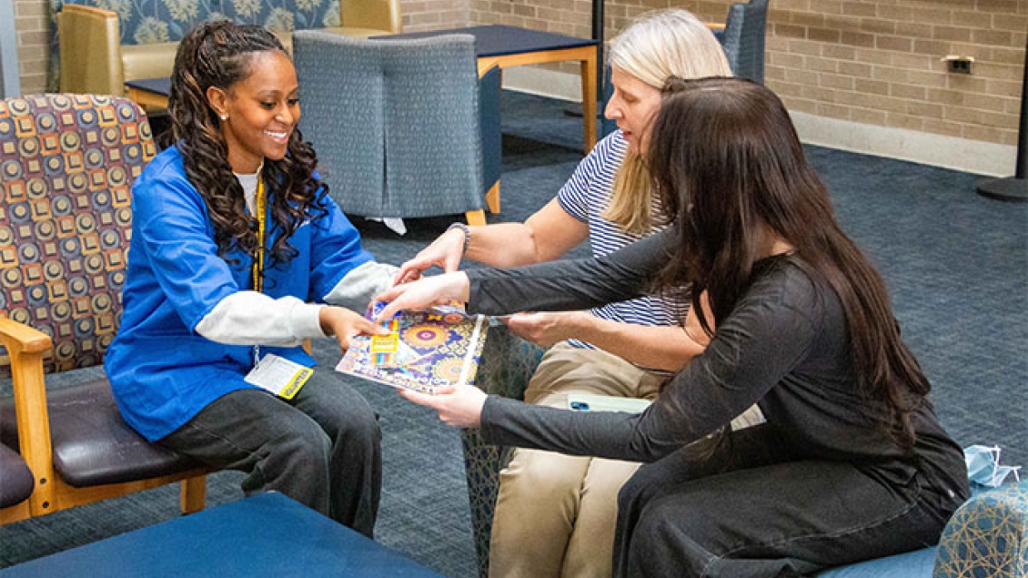 A smiling volunteer in a blue uniform hands a coloring book and crayons to two individuals sitting in a lounge area.