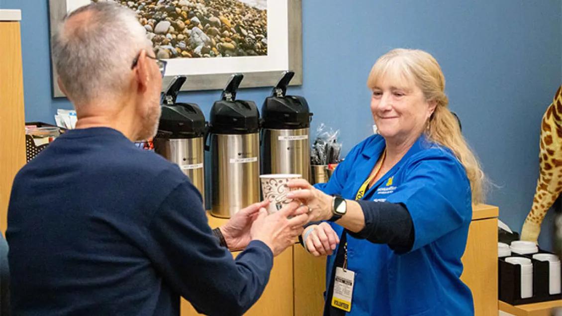 A volunteer wearing a blue University of Michigan shirt and yellow lanyard smiles while handing a cup of coffee to a man in a breakroom setting.