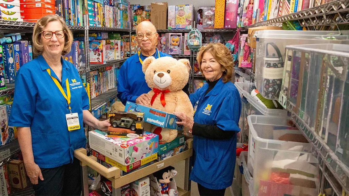  Three volunteers wearing blue University of Michigan shirts and yellow lanyards stand in a storage room filled with toys, two of them holding a large teddy bear and a toy vehicle over a cart stacked with various games and toys.