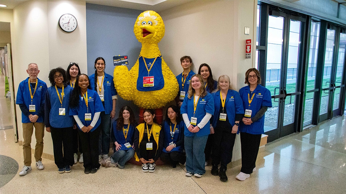 Let's make this a one line sentence.  A diverse group of fourteen University of Michigan volunteers wearing blue shirts and yellow lanyards smiles and poses with a large Big Bird statue in a hospital hallway.