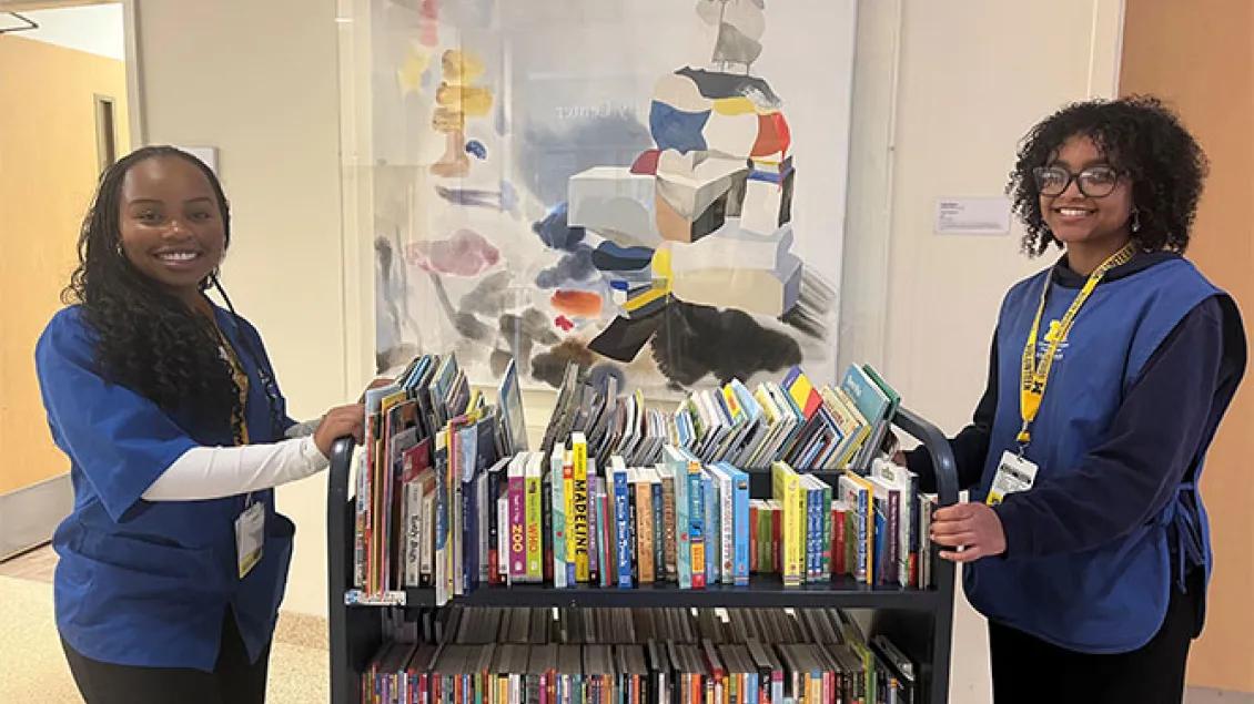  Two smiling volunteers wearing blue shirts and yellow lanyards stand on either side of a library cart filled with books in a hospital hallway.