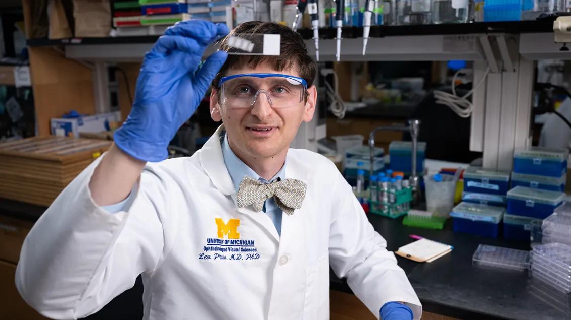 Lev Prasov, M.D., Ph.D., wearing a white coat, blue gloves, and safety glasses, views a microscope slide he is holding toward the viewer.