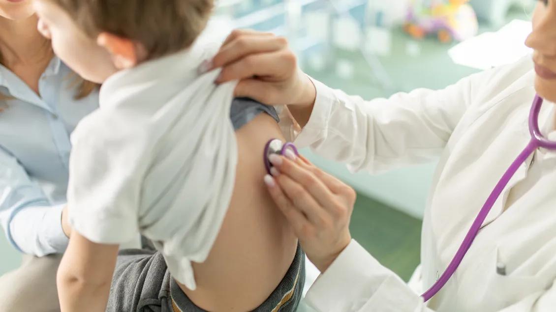A doctor holds a stethoscope to a young patient's back as a caregiver reassures him.