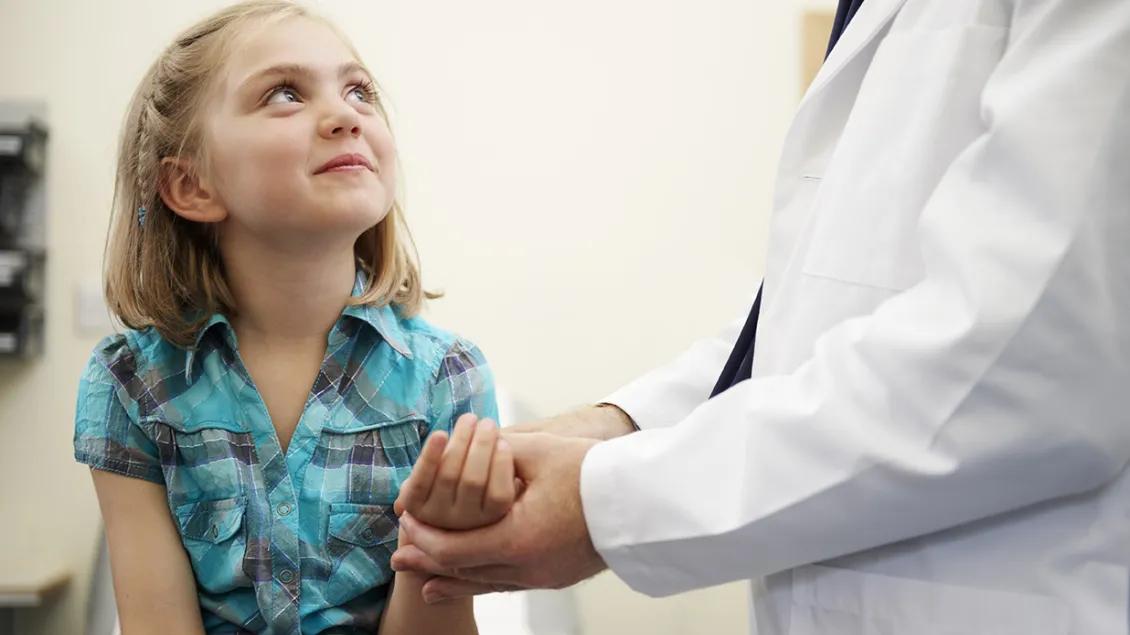 A young girl seated on an exam table looks up at a doctor who is examining her wrist.