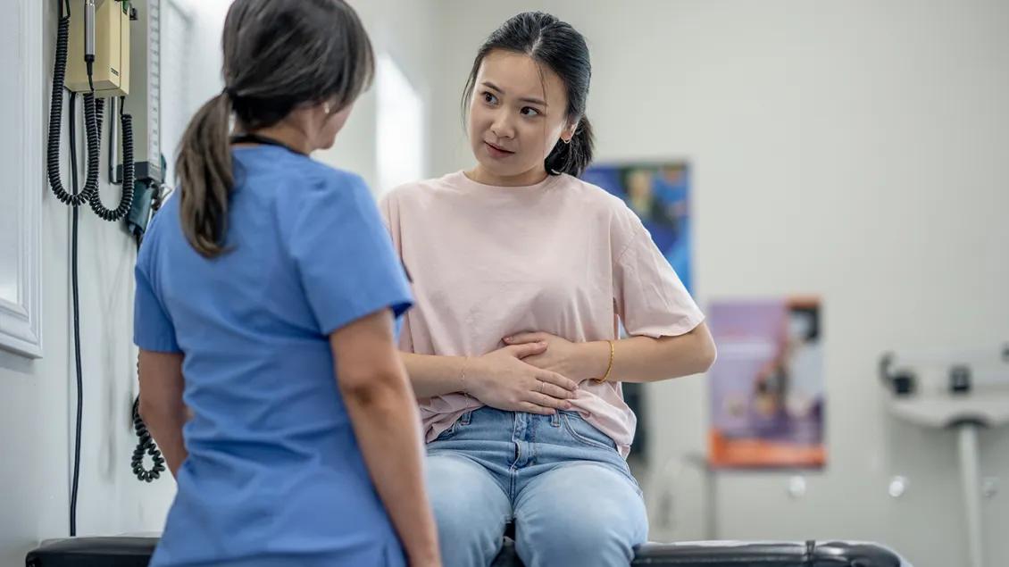 A patient seated on an exam table places her hands on her stomach as she talks to a health care professional.