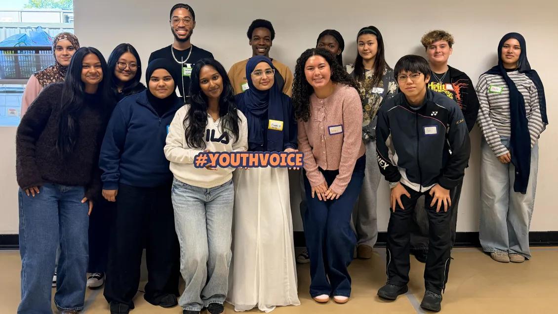 The 2026-2027 REACH group smiles in front of a wall. They hold a sign that says #YouthVoice.