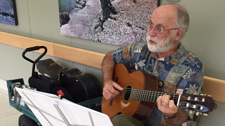 Older main with gray beard playing guitar in hospital hallway with art on wall behind